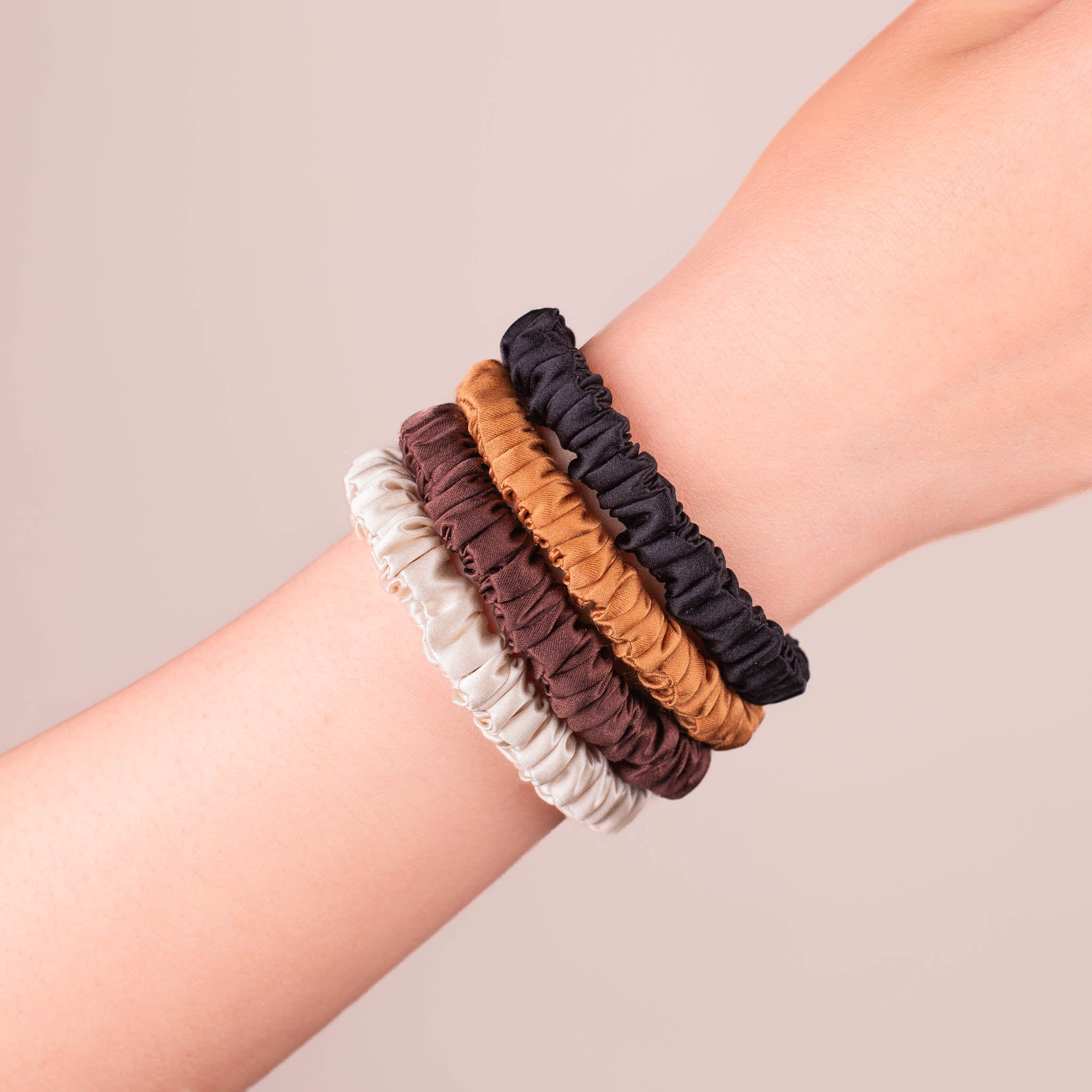 Set of 4 Skinny Silk Scrunchies in Warm Glow—Champagne, Dark Brown, Caramel, and Black—stacked on a wrist against a plain background.